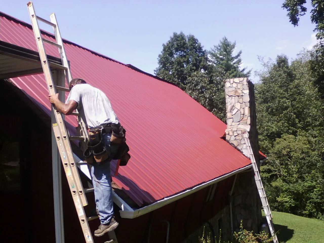 Roofer on a ladder beside a red metal roof and stone chimney — Soffit & Fascia Repair by Roofing Brothers in Beavercreek