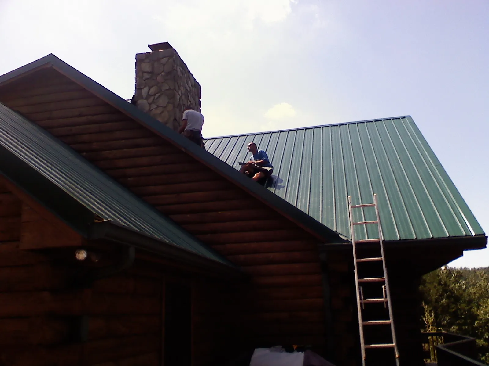 Roofers installing green metal panels beside a stone chimney on a log home in Beavercreek